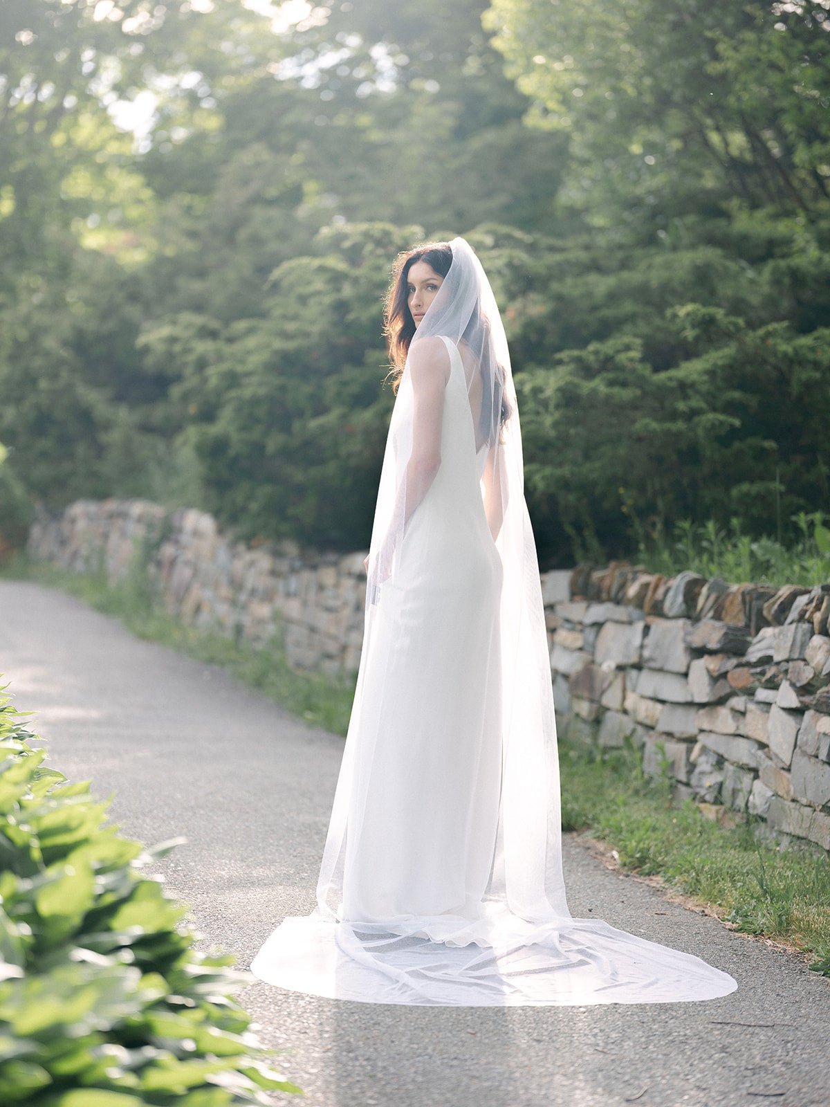 A bride is fully encased by her flowing bridal veil. Viewed from the back, the bride stands on a path amongst greenery while looking back.