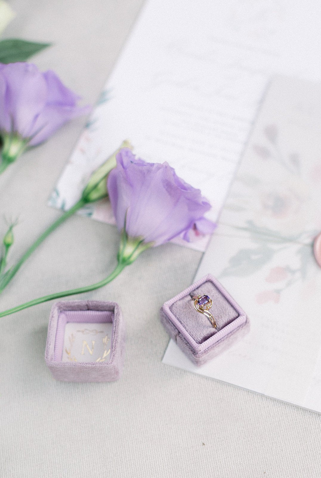 A distanced shot of a Diamond and Amethyst Vintage Engagement Ring in 10k Yellow Gold in a purple ring box on white background with purple flowers. 