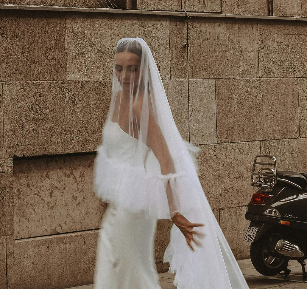 A bride in sleek white bridal gown draped in a sheer tulle wedding veil with sandy brick building and back end of a scooter as background.