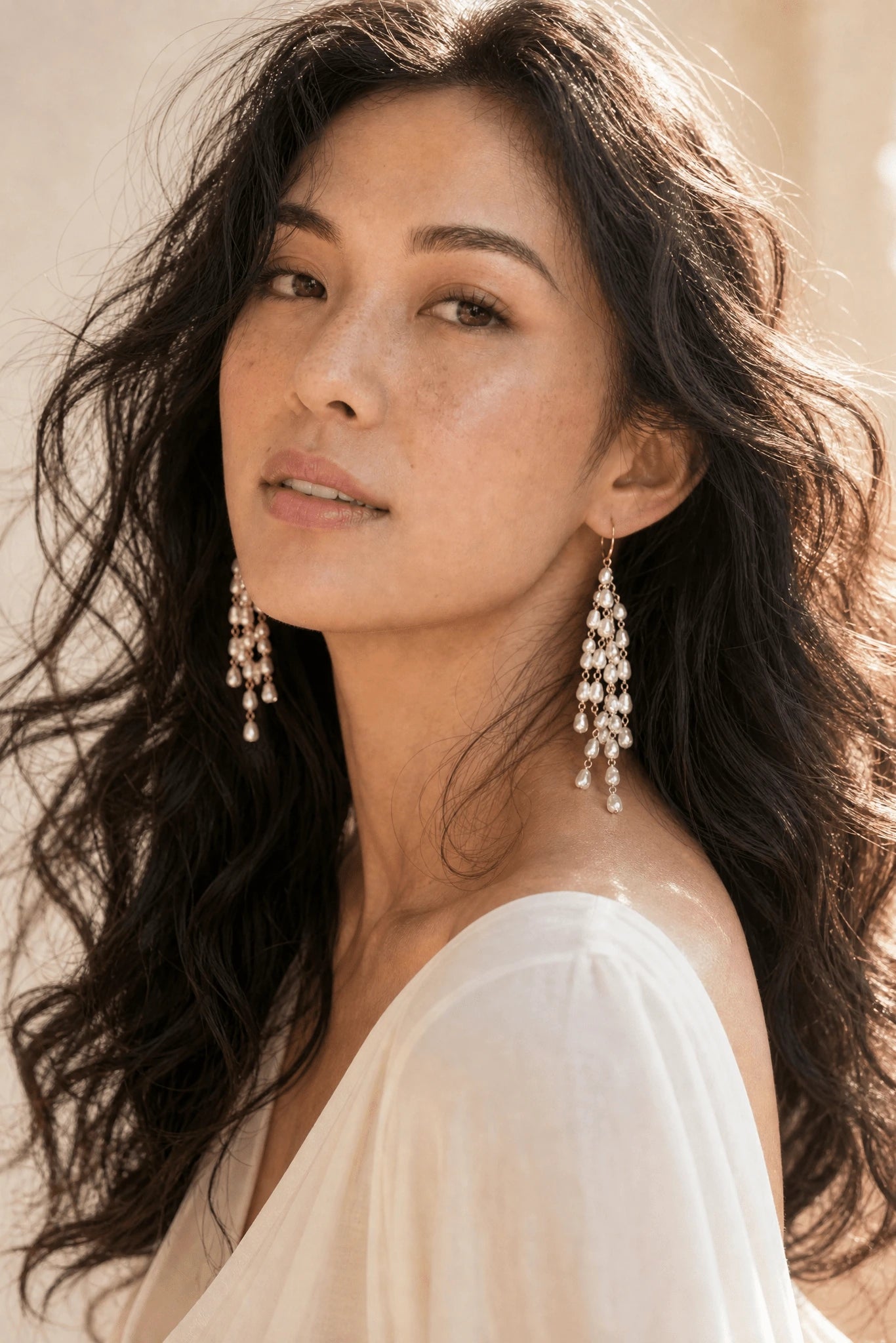 Filipino woman with wavy dark hair in white wearing large freshwater pearl tassel statement earrings against neutral background. Handmade pearl earrings by Canadian jewellery designer