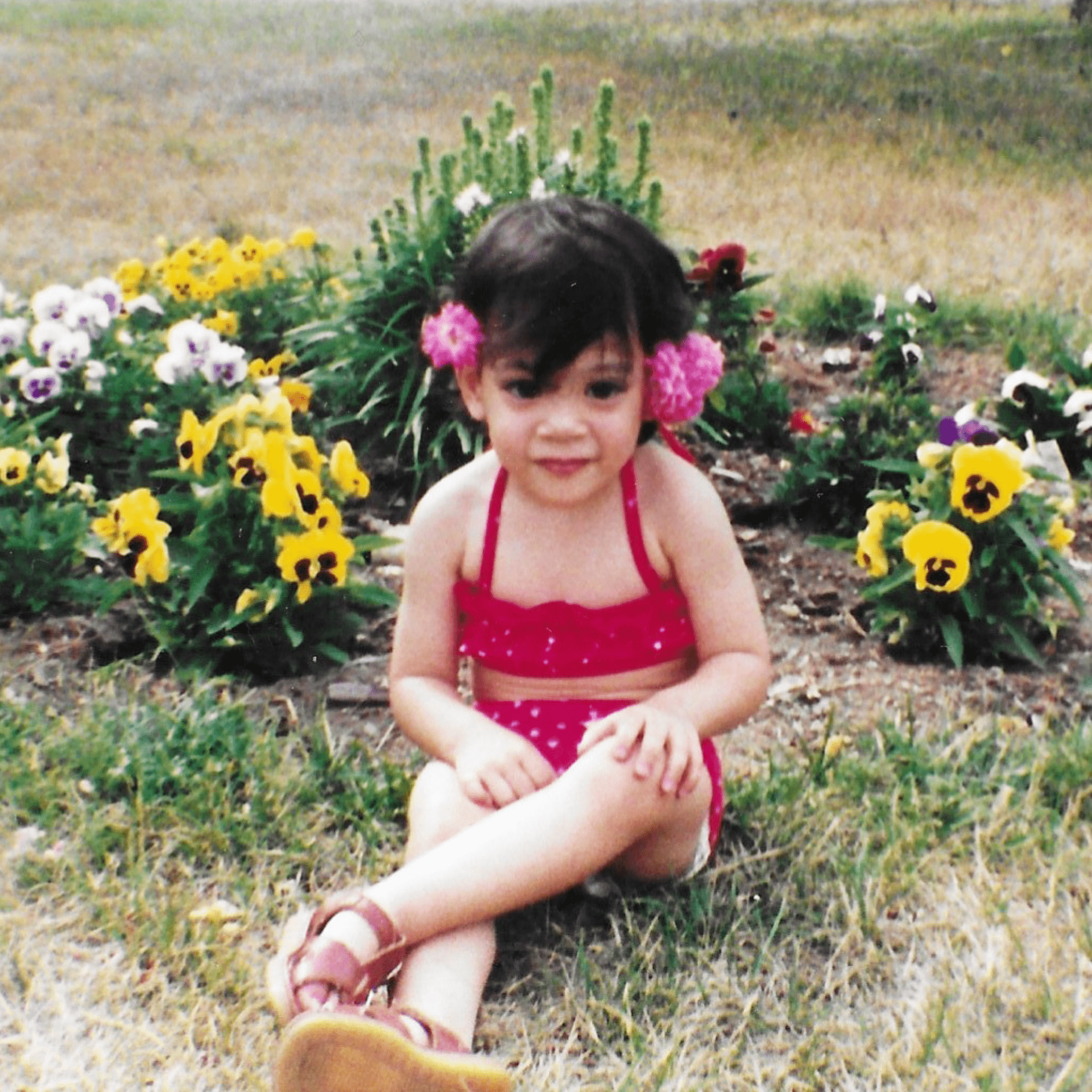 Child in a red outfit sitting among flowers in a garden.