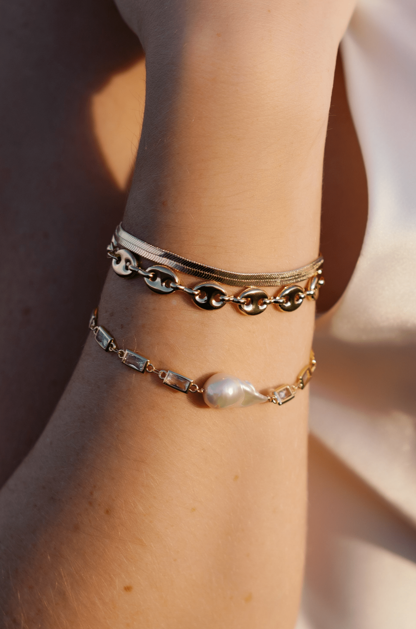 A stack of 18k gold-filled bracelets that include a herringbone chain bracelet, puffy mariner link bracelet, and crystal link with baroque pearl bracelet.